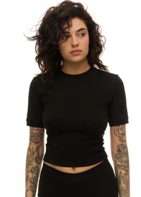 Woman with dark curly hair in a black cropped t-shirt, looking to the left; tattoos on both arms visible against a white background.