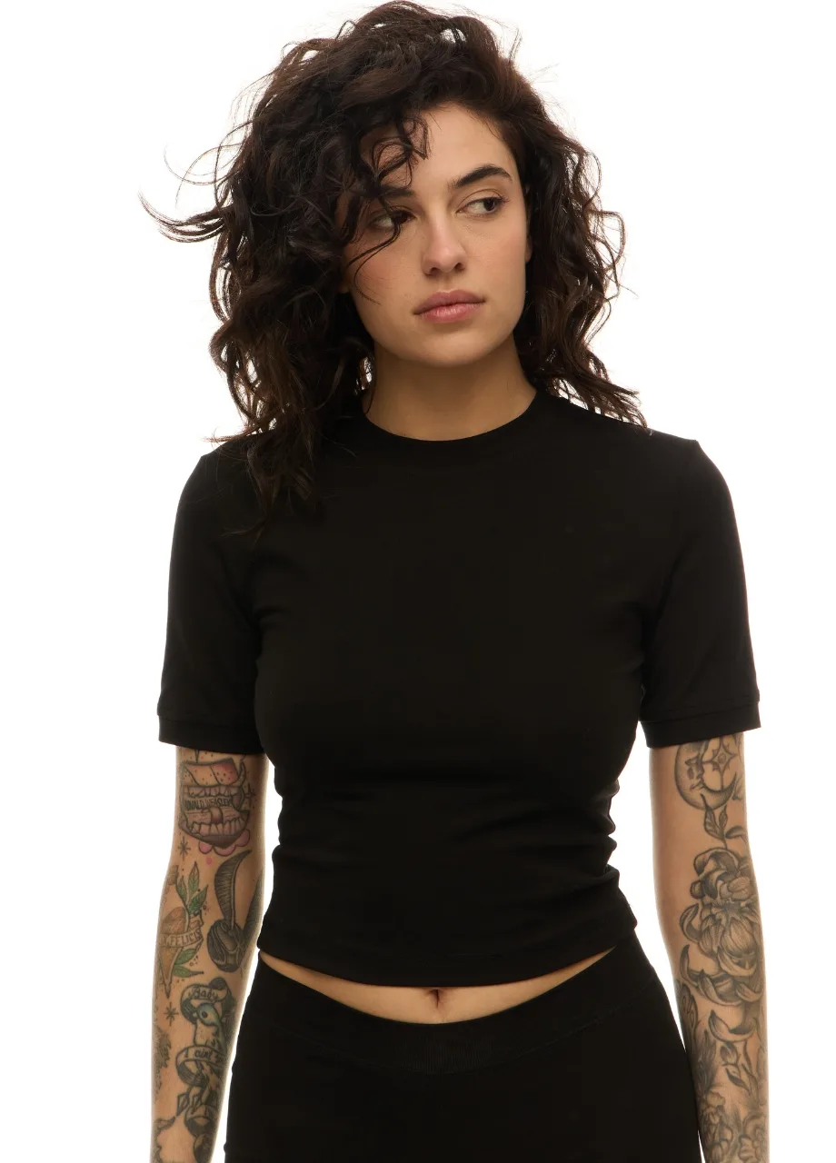 1 › Str/Cd Wear Woman with dark curly hair in a black cropped t-shirt, looking to the left; tattoos on both arms visible against a white background.