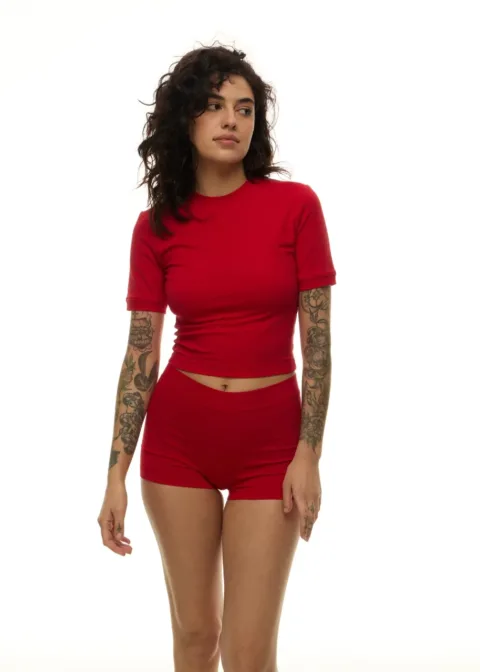 Woman with curly dark hair wearing a red cropped top and red shorts, tattoos on both arms, standing against a white background.