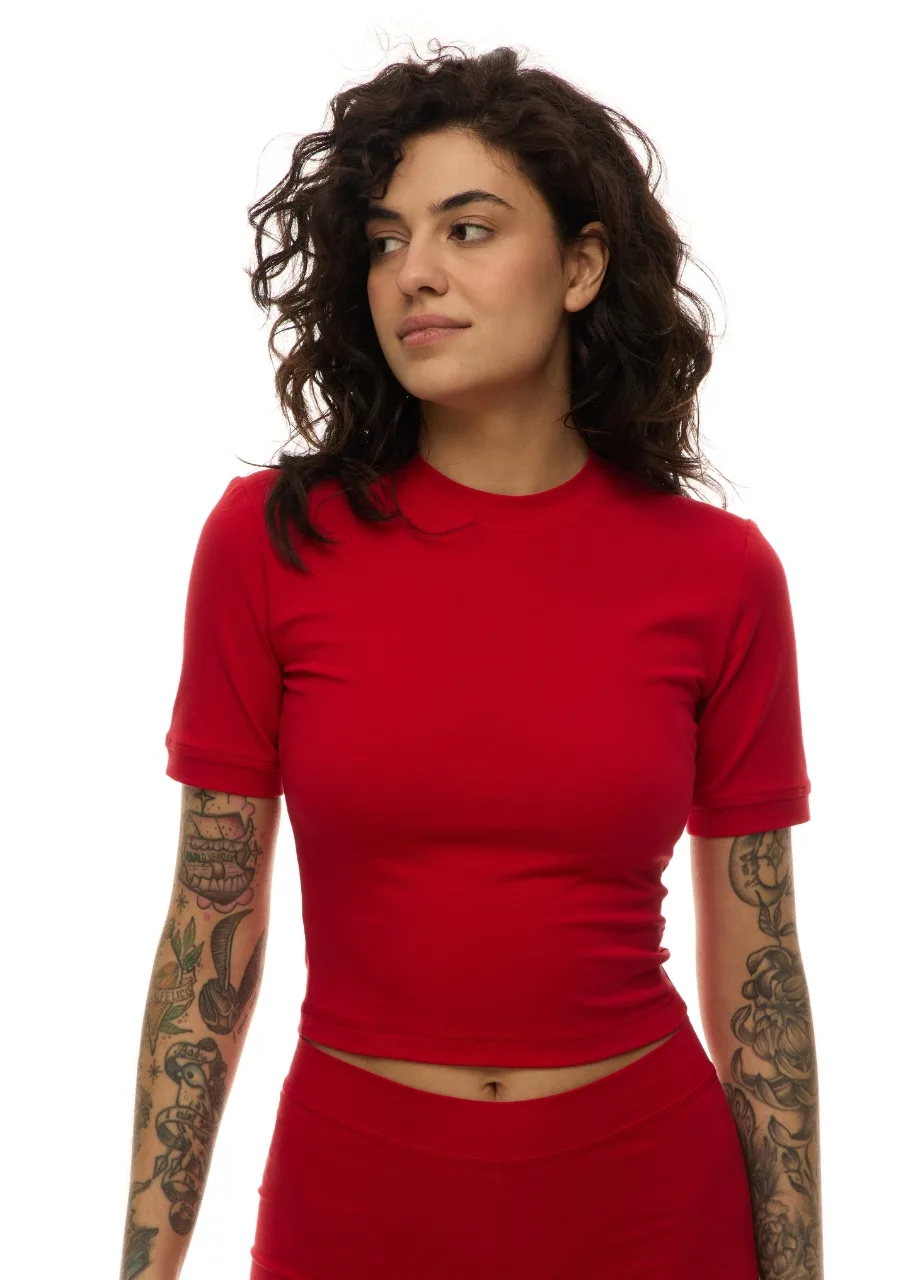 16 › Str/Cd Wear Woman with curly dark hair wearing a fitted red short-sleeve top and matching red pants, looking to the left, showing tattoos on both arms.