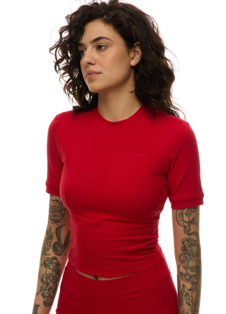 17 › Str/Cd Wear Profile view of a tattooed woman in a bright red fitted dress against a white background, with dark curly hair.