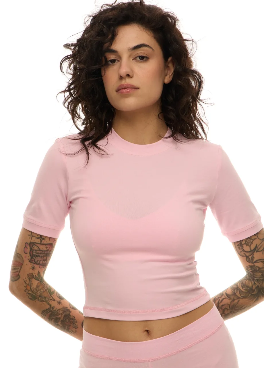 19 › Str/Cd Wear Woman with dark wavy hair wearing a pale pink cropped T-shirt and matching high-waisted shorts, standing with hands on hips, arms tattooed.