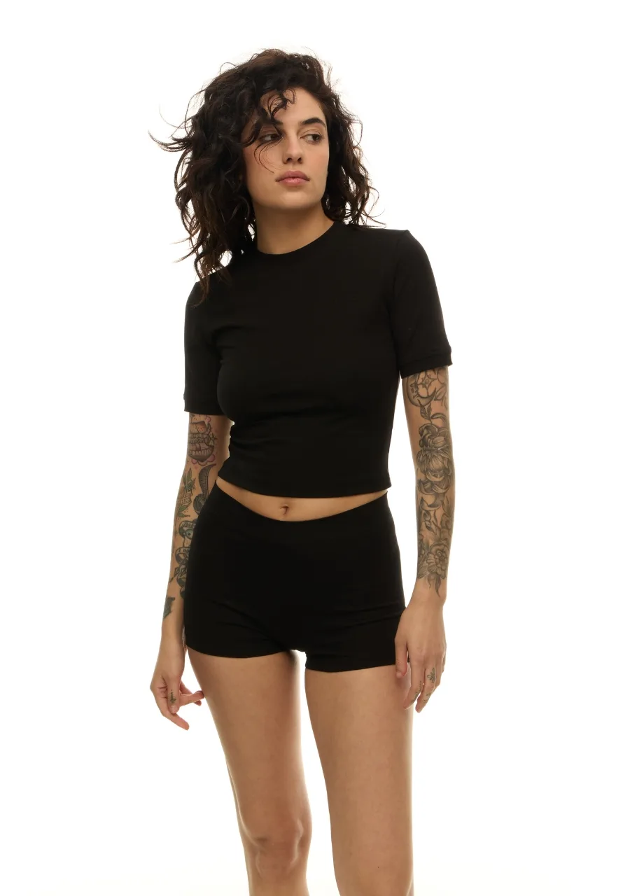 2 › Str/Cd Wear Woman wearing a black cropped t-shirt and matching black shorts, standing with arms at her sides, showing tattooed arms against a plain white background.