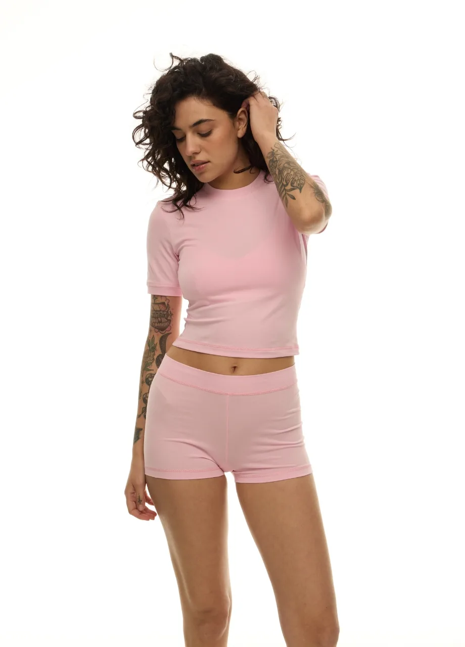 20 › Str/Cd Wear Woman wearing a light pink cropped top and matching shorts, standing against a white background with one hand in her hair and visible arm tattoos.