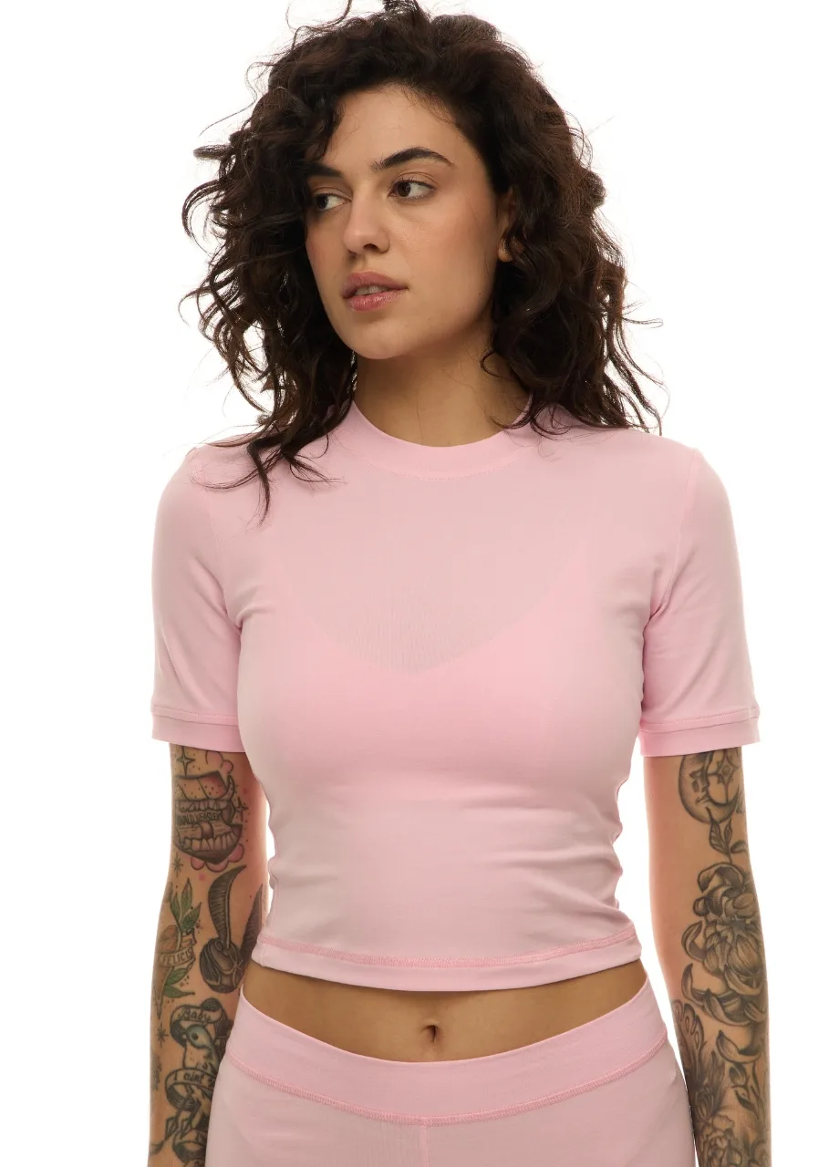 21 › Str/Cd Wear Woman with dark curly hair wearing a light pink cropped t-shirt; visible tattoos on both forearms.