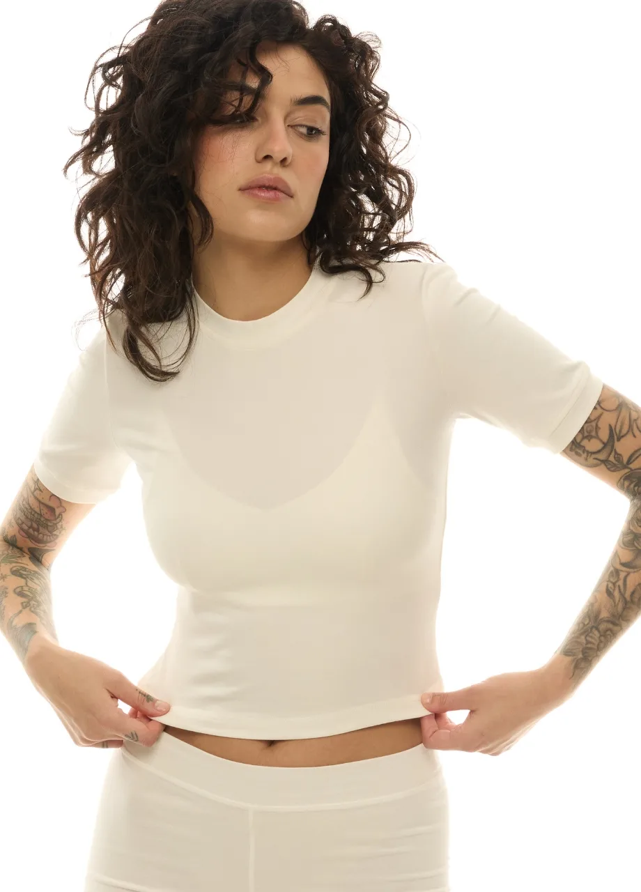 5 › Str/Cd Wear Woman with dark, curly hair wearing a white tee and white bottoms, lifting the hem to reveal her midriff and tattoos on her forearms.