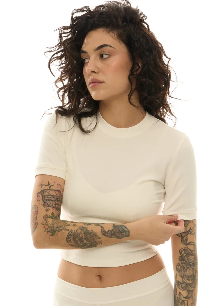 6 › Str/Cd Wear Woman with dark, curly hair wearing a white cropped top, arms crossed, looking to the left; visible tattoos on both forearms.