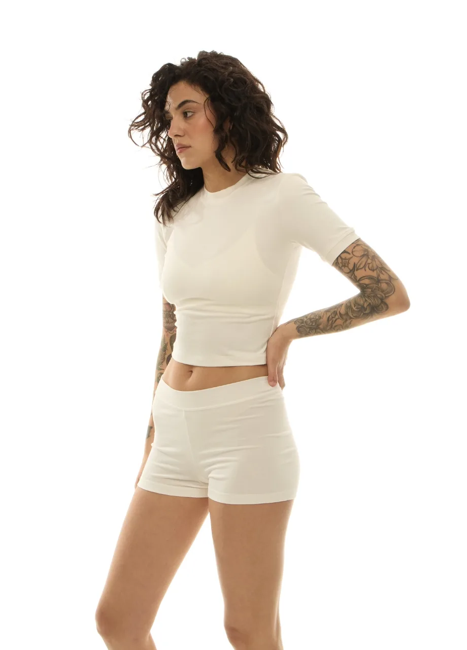 7 › Str/Cd Wear Woman with curly dark hair in a white crop top and matching shorts, standing in profile with her left hand on her hip (arm tattoos visible).