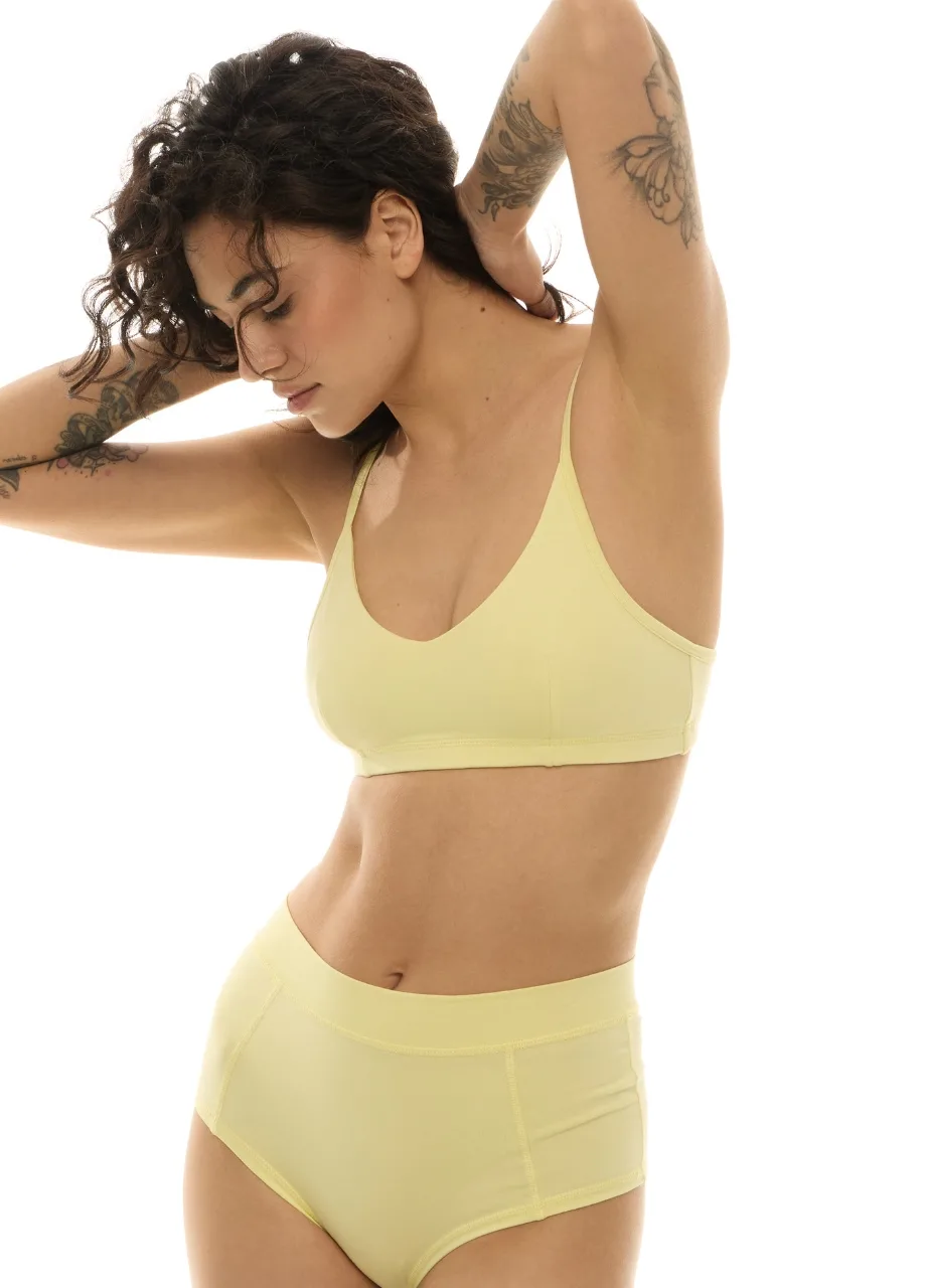 WP611222 › Str/Cd Wear Woman with short dark hair in a pale yellow bra and high-waisted underwear, arms raised behind her head, tattoos on her left arm visible (profile view).
