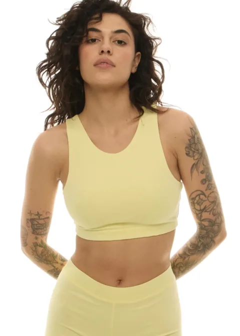 Woman wearing a pale yellow sports bra and matching high-waisted leggings, standing with hands behind her back, tattoos visible on arms, neutral expression.