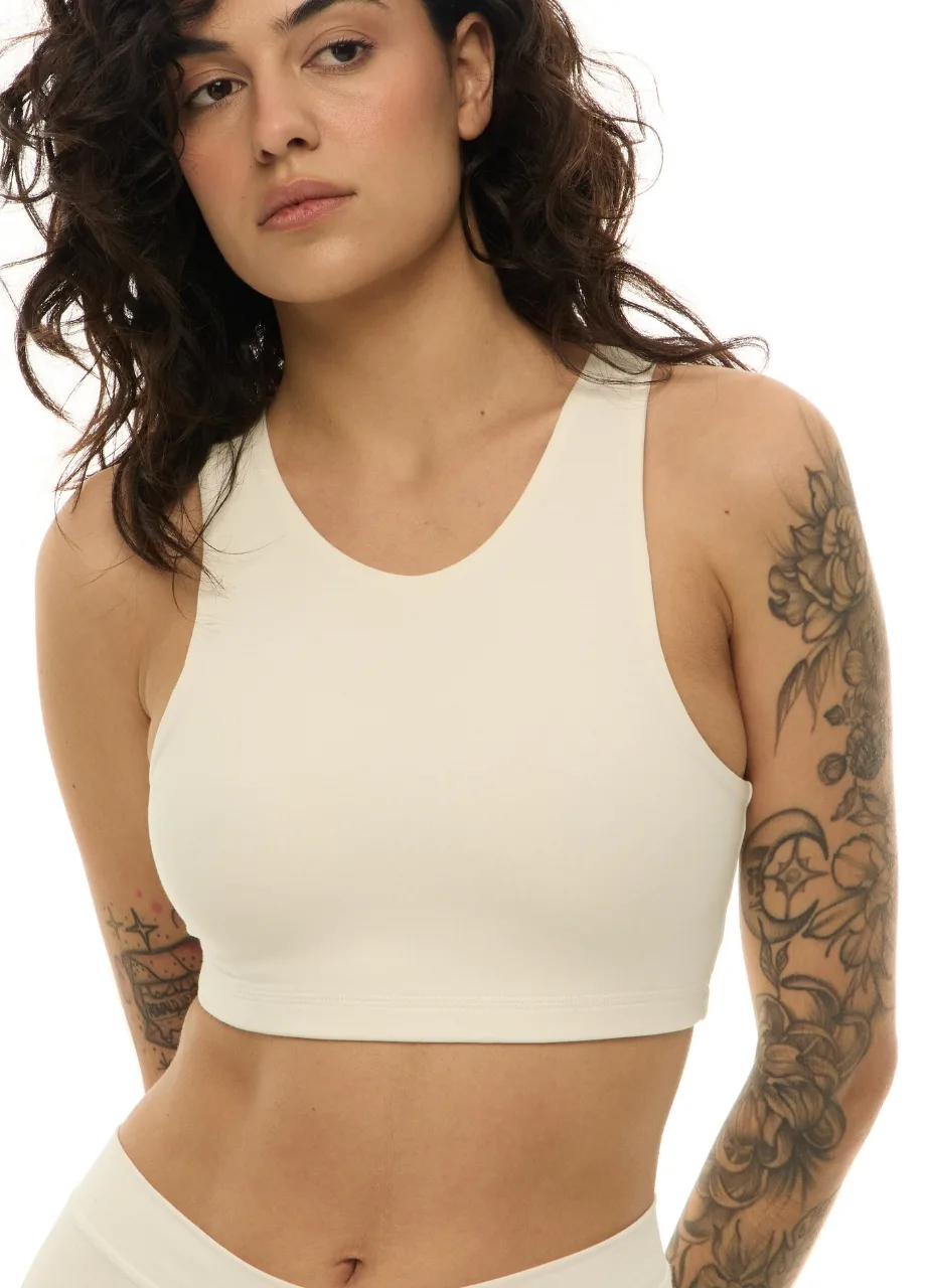 WS50953 › Str/Cd Wear Woman with wavy dark hair wearing a cream sports bra, visible tattoos along her right arm, torso shot.
