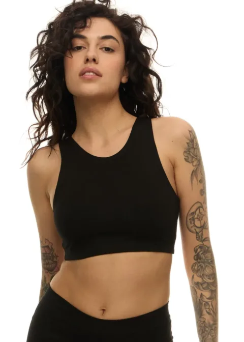 Woman with curly dark hair wearing a black sports bra, exposing tattoos on both arms, posing against a white background.