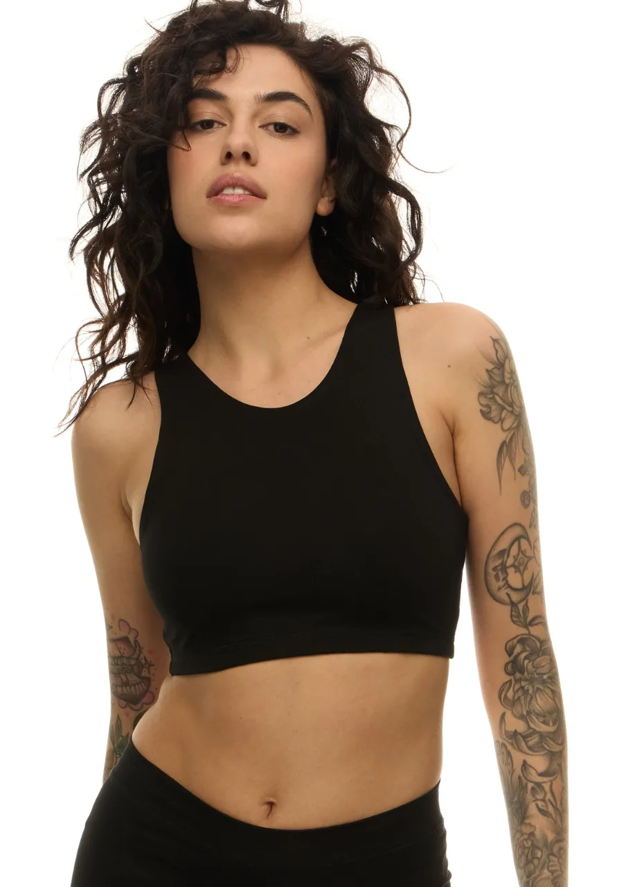 WS50956 › Str/Cd Wear Woman with curly dark hair wearing a black sports bra, exposing tattoos on both arms, posing against a white background.