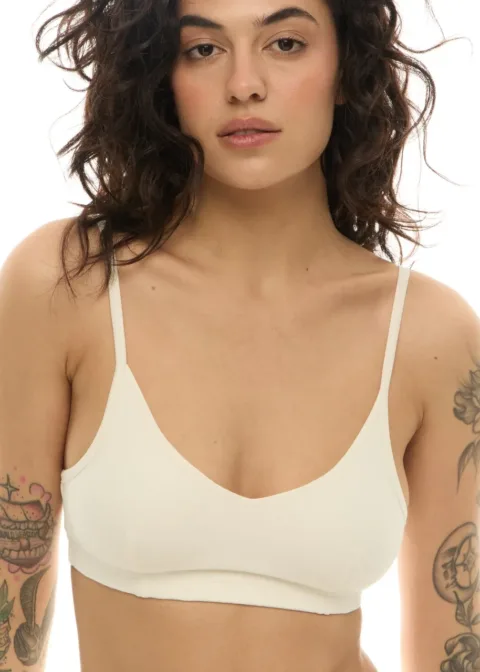 Model wearing a white bralette facing forward with visible arm tattoos and neutral expression.