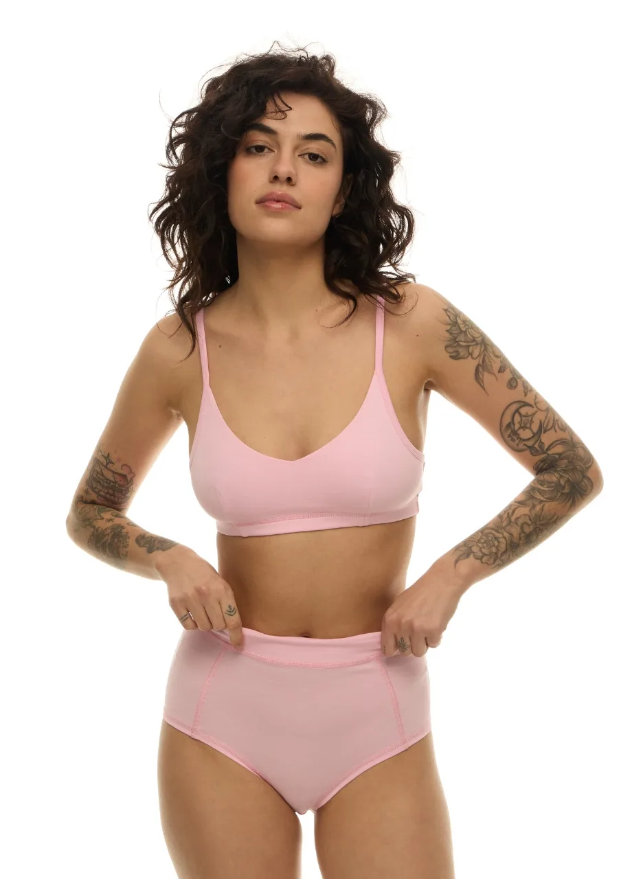 WS511111 › Str/Cd Wear Model wearing a light pink bralette and high-waisted panties, standing with hands at her waistband against a white background, showing tattoos on arms.