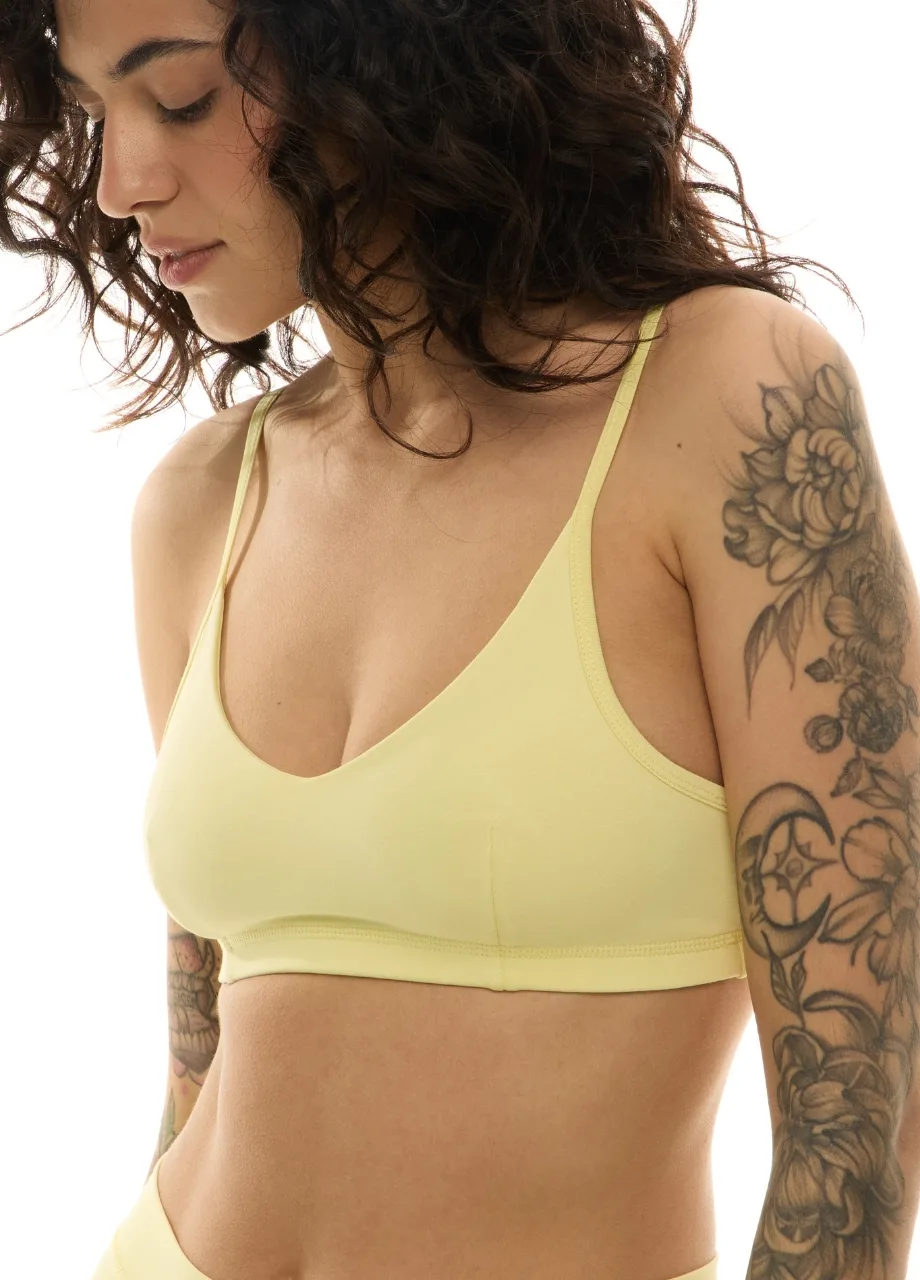 WS511121 › Str/Cd Wear Side-profile of a woman wearing a pale yellow bralette, showcasing her shoulder-length curly hair and floral tattoo on her arm.