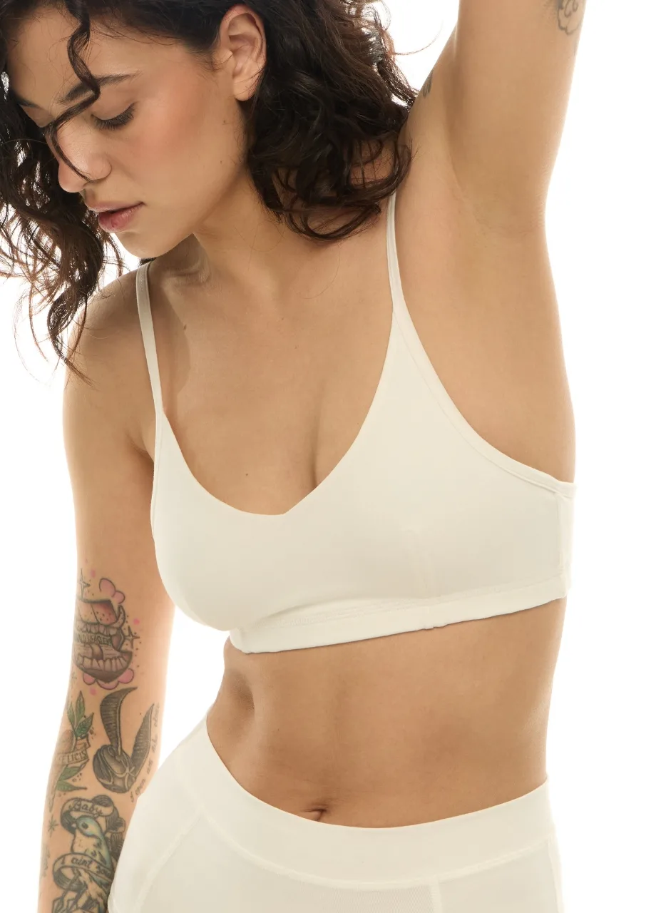WS51113 › Str/Cd Wear Woman in a cream sports bra with her left arm raised, tattoos visible on her left forearm in a side profile.