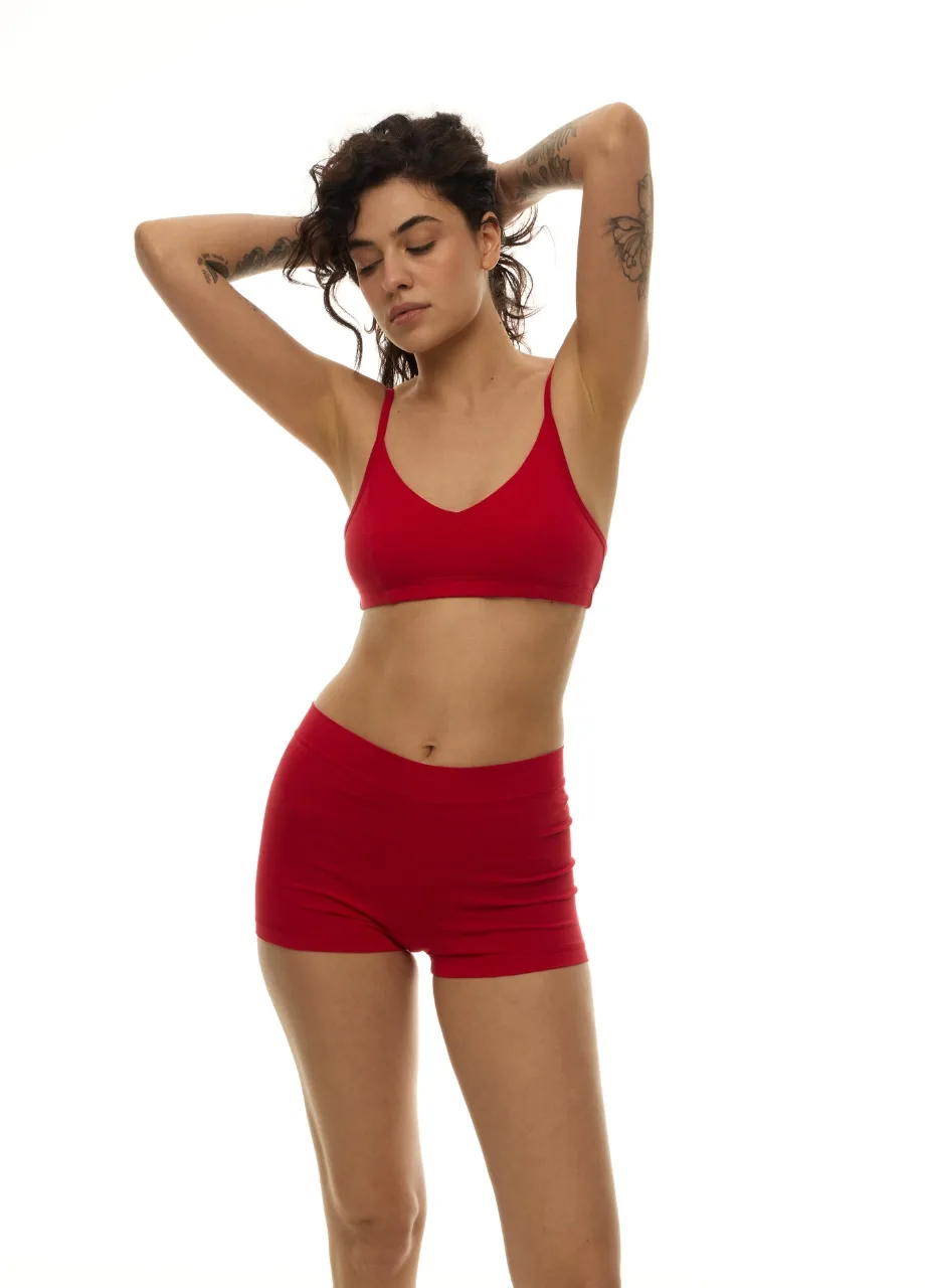 WU1313016 › Str/Cd Wear Woman in a red sports bra and matching shorts poses with arms raised behind her head against a white background.