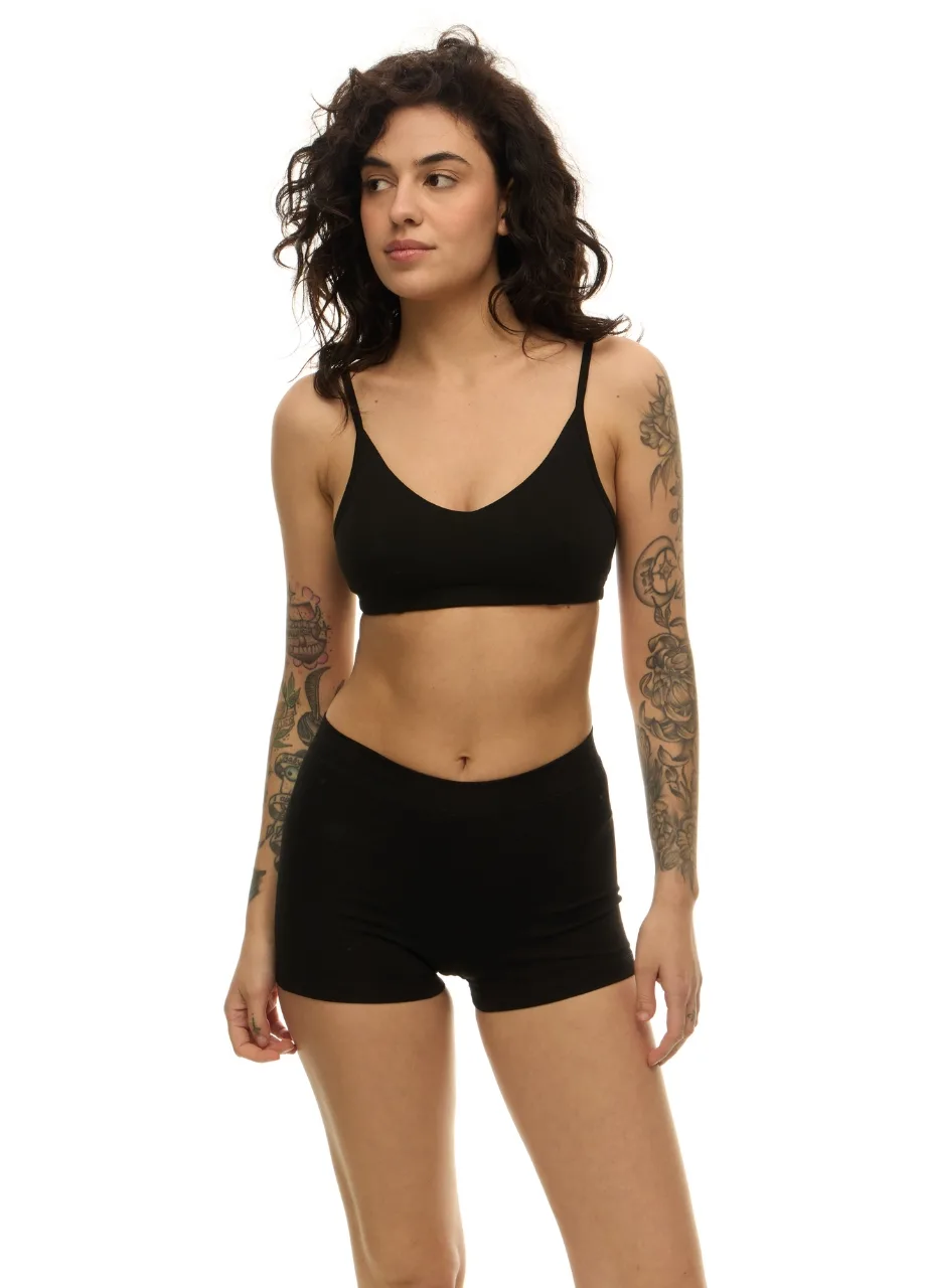 WU131308 › Str/Cd Wear Woman with curly dark hair wearing a black sports bra and black shorts, standing against a white background, tattoos visible on both arms.