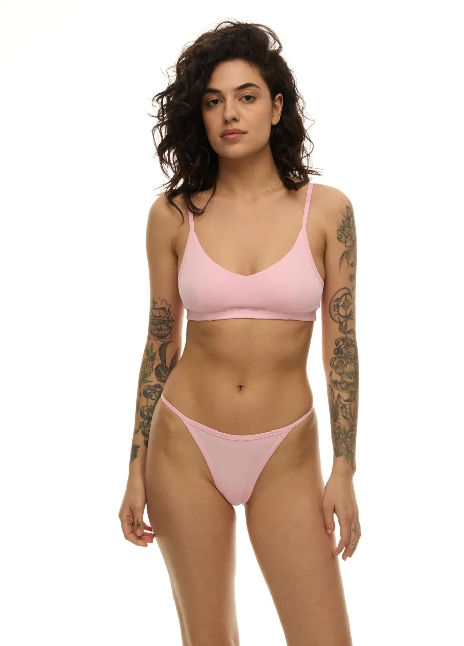 WU1314114 › Str/Cd Wear Woman in light pink bra and thong standing against a white background, facing the camera, with tattoos on both arms.