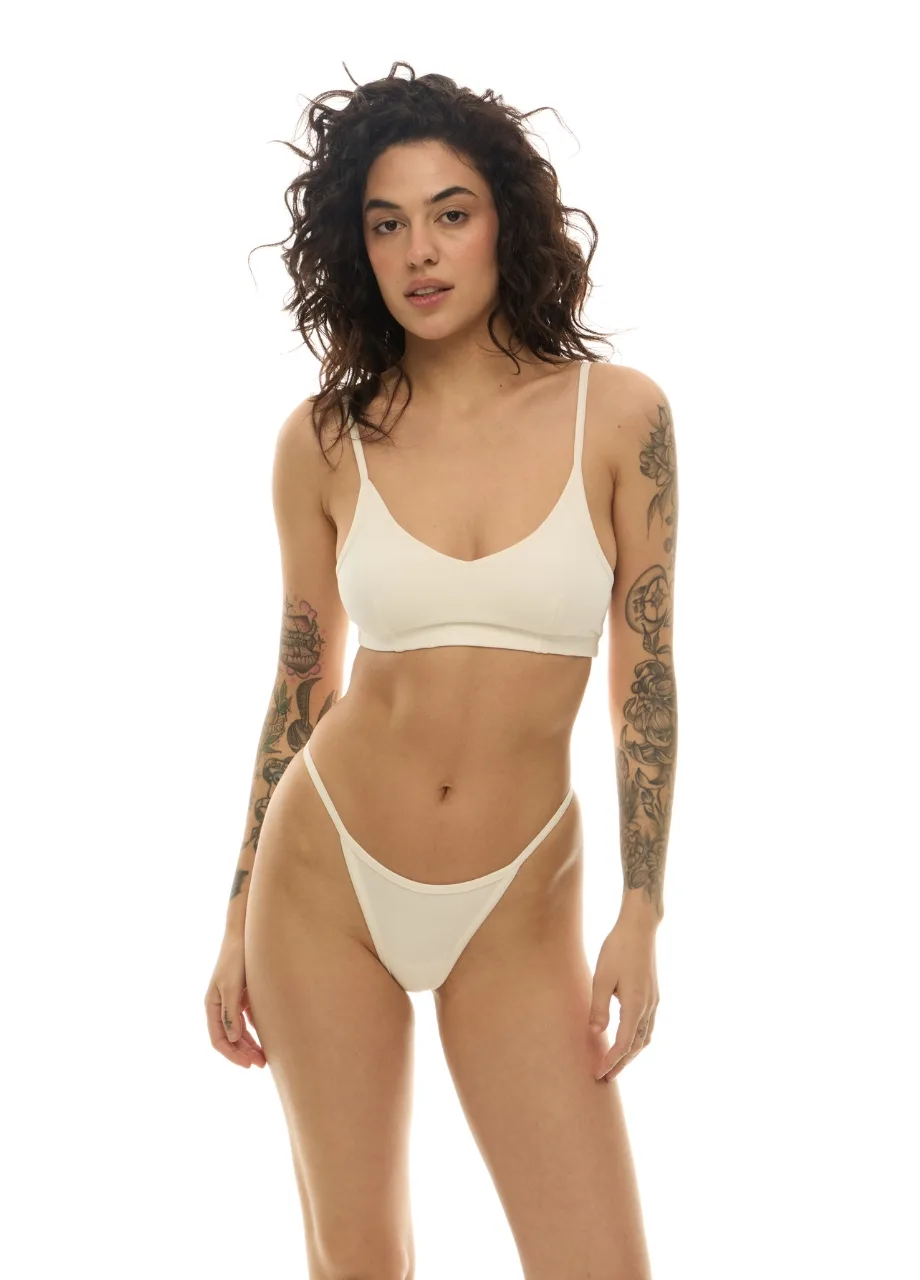 WU131412 › Str/Cd Wear Woman modeling cream-colored lingerie—a bralette and high-cut thong—with tattoos on her arms, standing against a white backdrop.