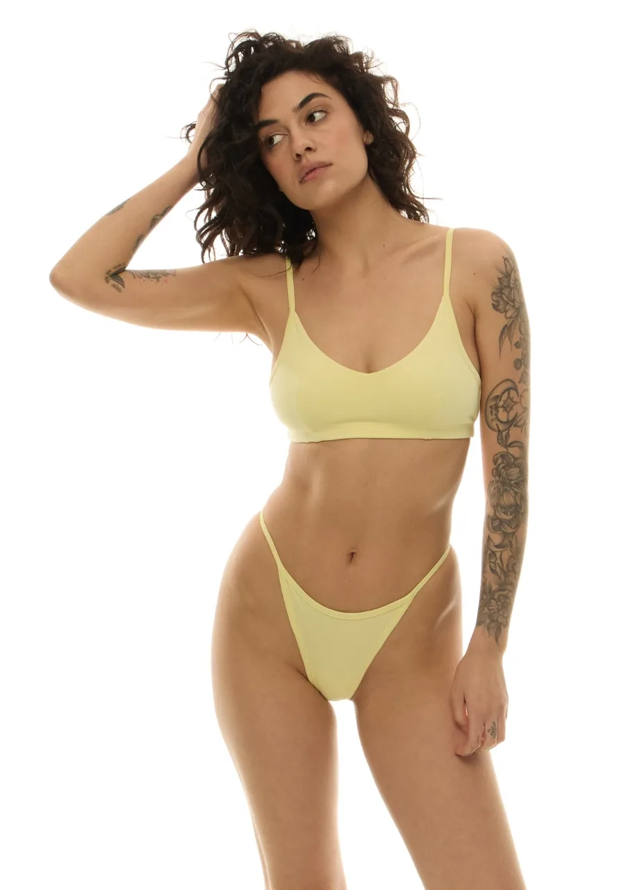 WU1314122 › Str/Cd Wear Woman in pale yellow bikini set (bra and high-cut bottoms) poses with one hand in her hair, visible tattoos on her left arm and side.