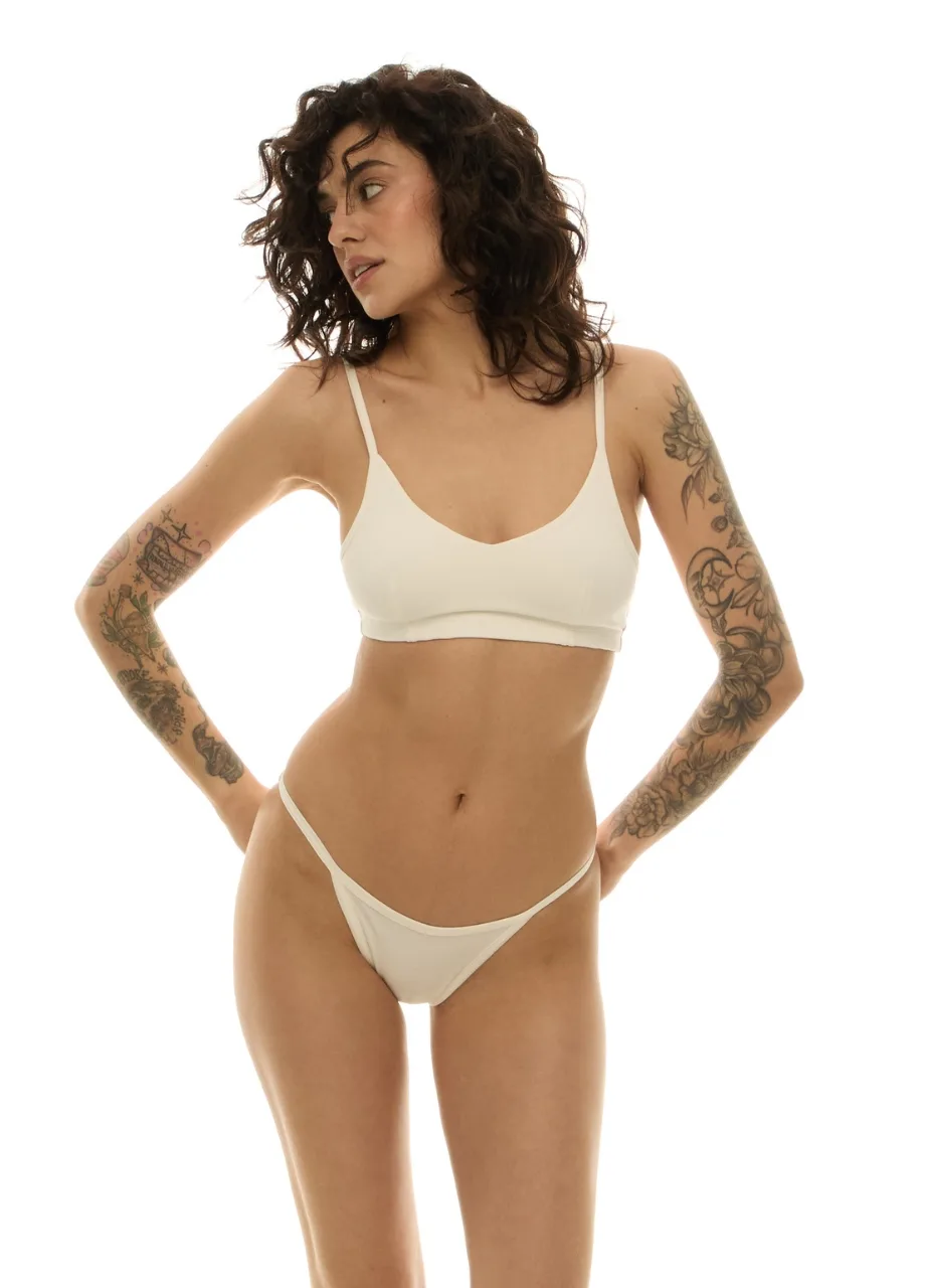 WU131425 › Str/Cd Wear Woman with curly short hair wearing a beige bra and matching thong, standing with hands on hips and tattoos on arms, against a white background.