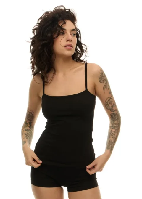 Woman with curly dark hair wearing a black tank top and black shorts, standing with hands at hips, showing full sleeves of tattoos on both arms.