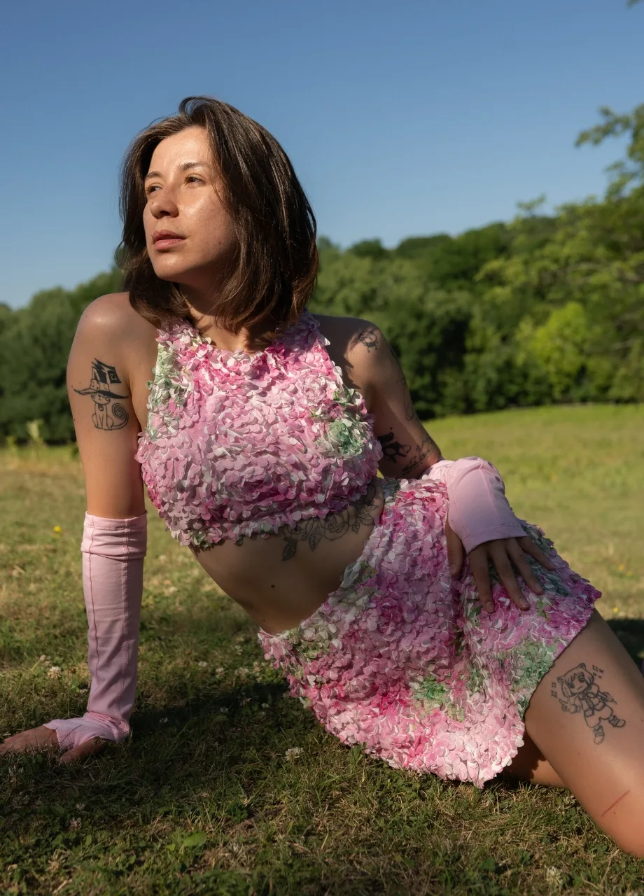 3 › Str/Cd Wear Woman with short brown hair sits on grass in a pink floral two-piece outfit, outdoors on a sunny day, tattoos visible on arms and leg.
