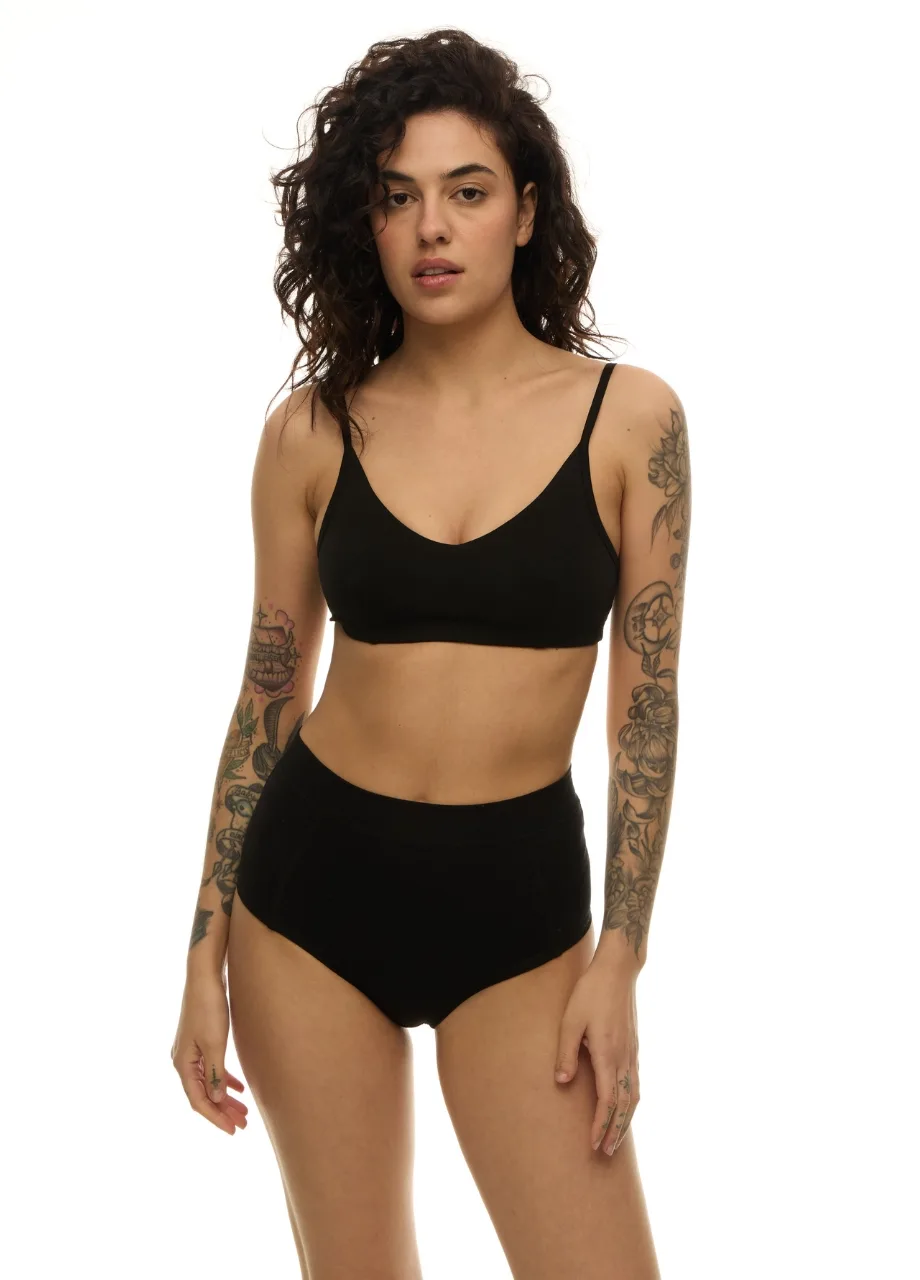 3 › Str/Cd Wear Woman wearing a black bralette and high‑waisted briefs, standing against a white background, showing tattoos on both arms.