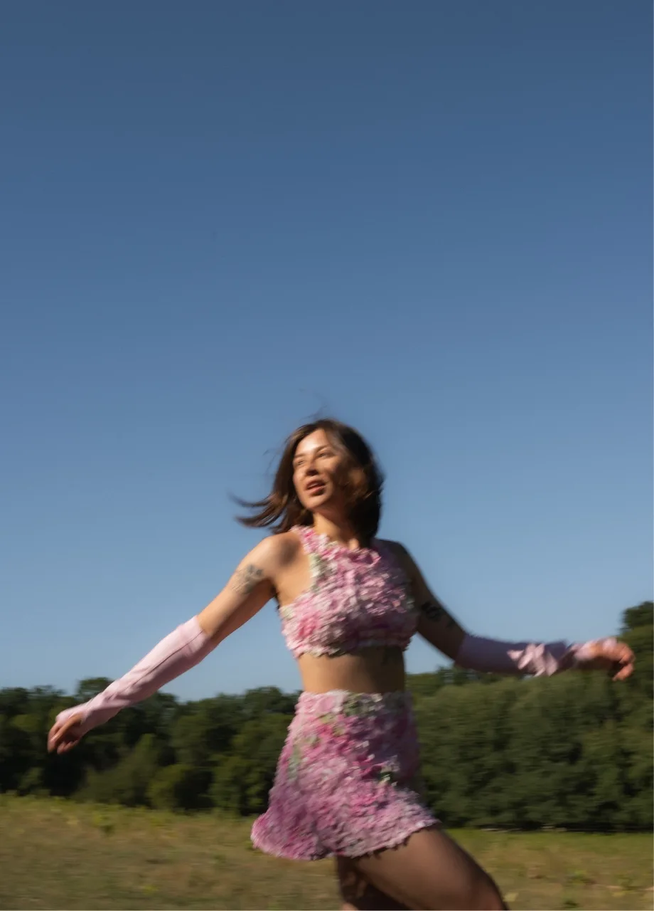3 › Str/Cd Wear Woman dancing in a pink floral two-piece outfit in an open field under a blue sky, motion blurred by movement.