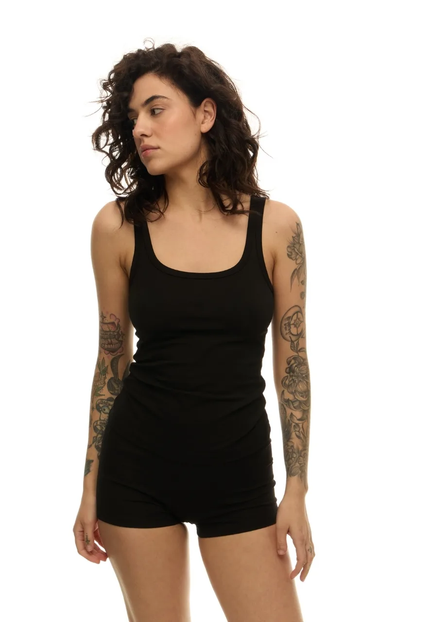 32 › Str/Cd Wear Woman with dark curly hair wearing a black tank top and black shorts, facing left with arms relaxed, tattoos visible on both arms.