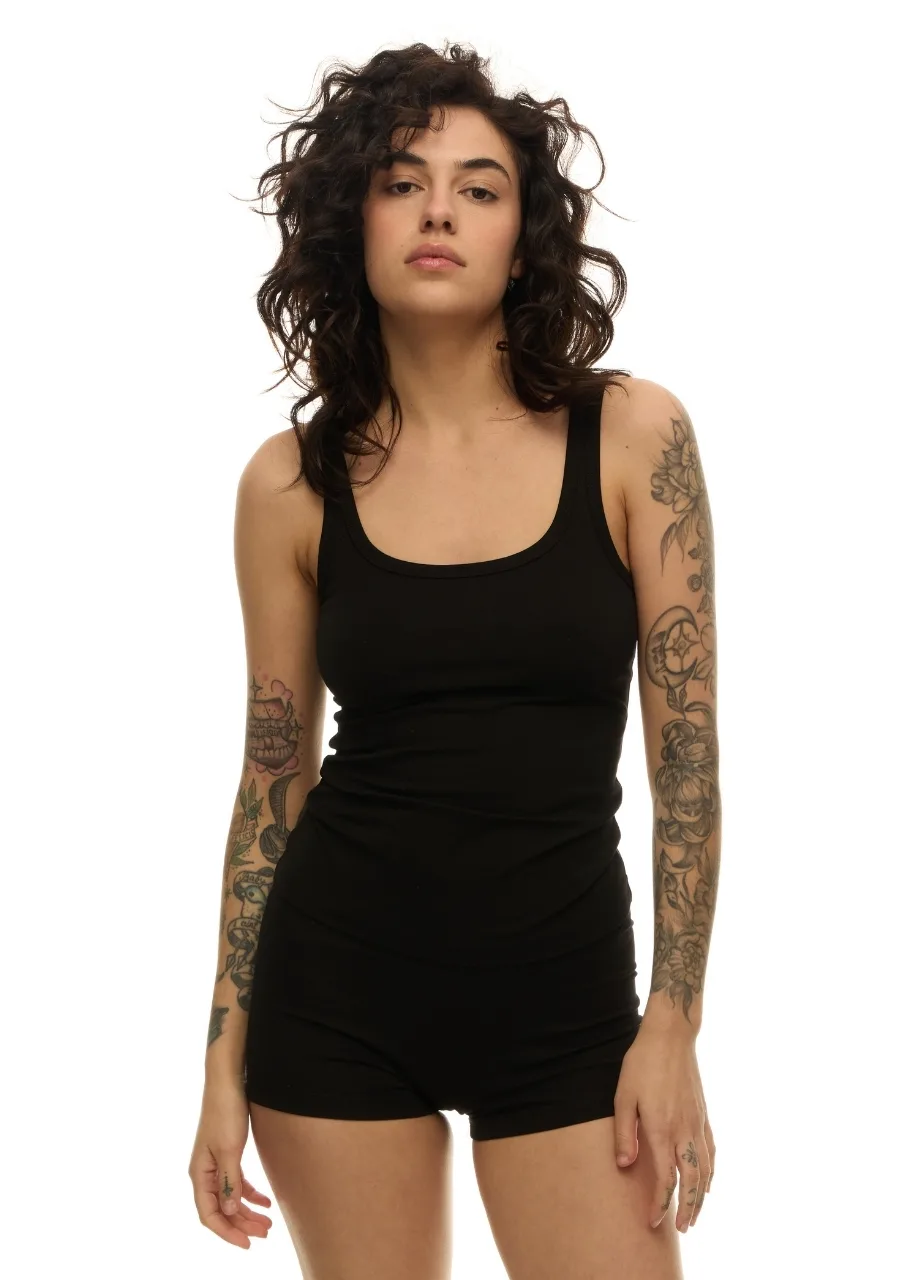 33 › Str/Cd Wear Woman with dark wavy hair wearing a black tank top and black shorts, standing against a white background with tattoos on her arms.