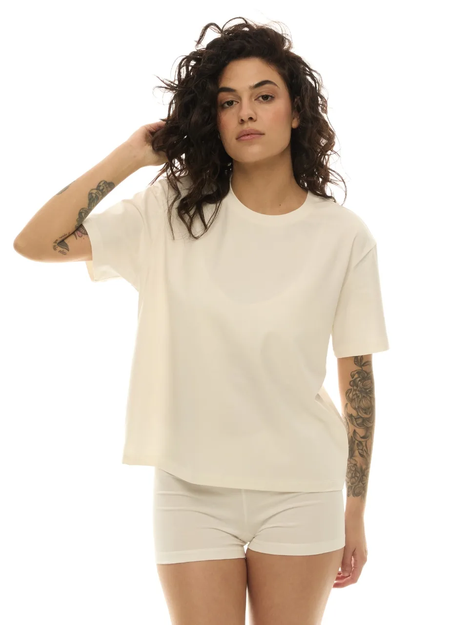 36 › Str/Cd Wear Woman with dark curly hair wearing an oversized cream T-shirt and matching shorts, showing tattooed arms and relaxed pose.