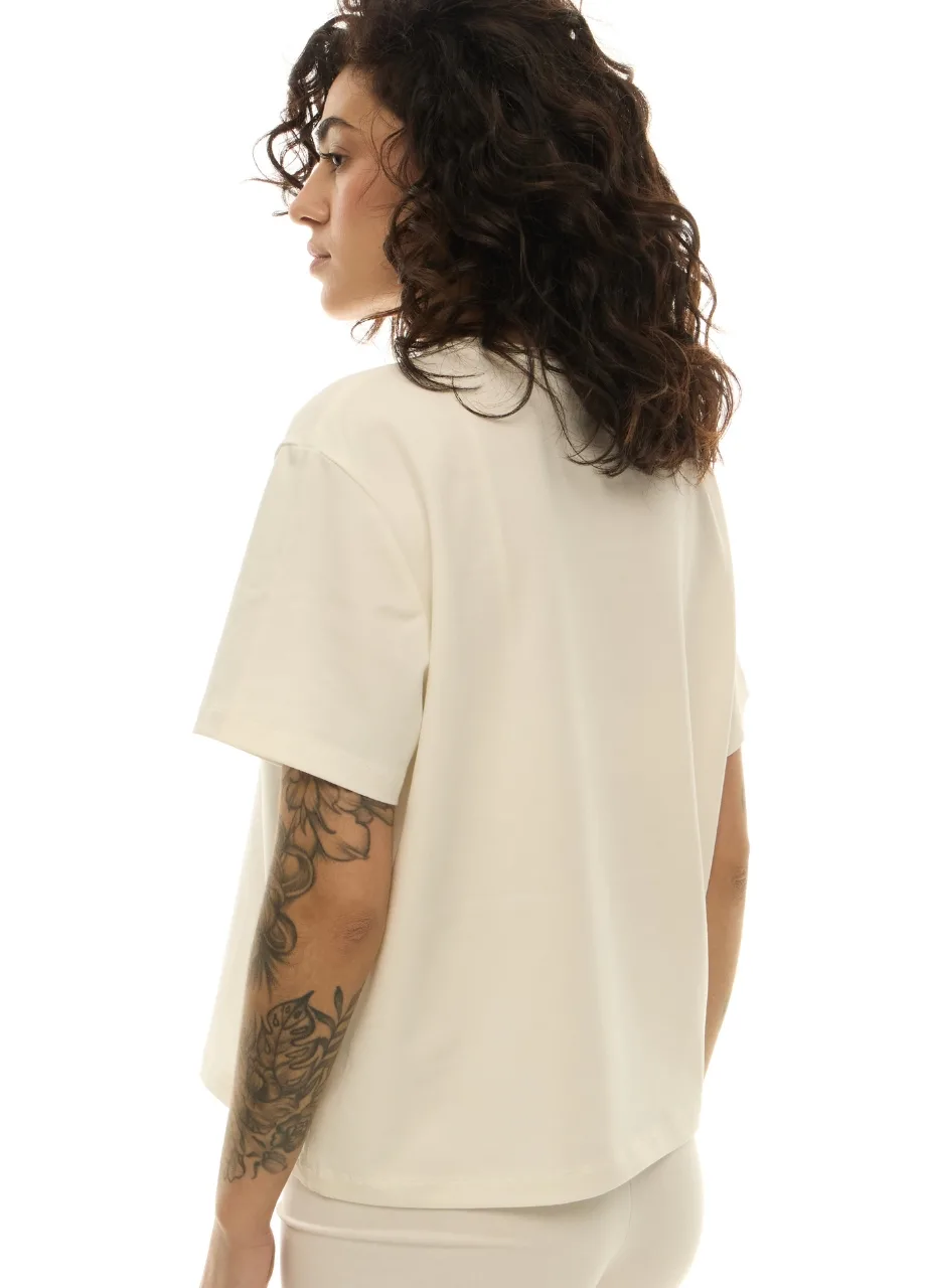 37 › Str/Cd Wear Back view of a person with curly dark hair wearing a loose cream T-shirt; a tattoo runs along their forearm.
