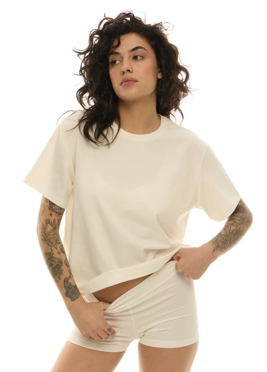 39 › Str/Cd Wear Woman with curly dark hair wearing an oversized cream T-shirt and matching shorts, posing with her hand at the hem.