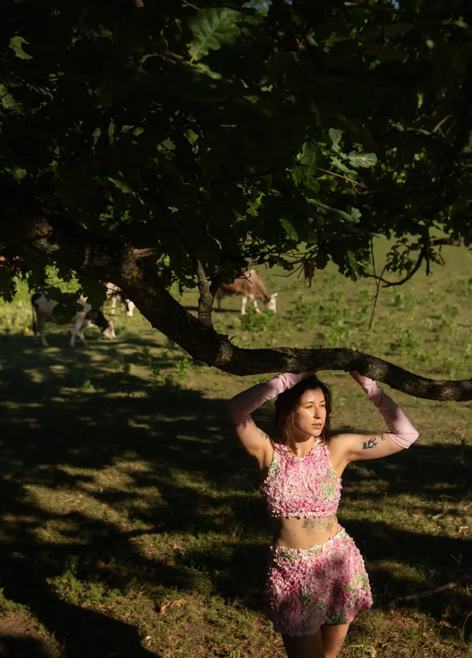 4 › Str/Cd Wear Woman in a pink floral outfit stands under a tree, holding a thick branch above her head in a sunlit field.