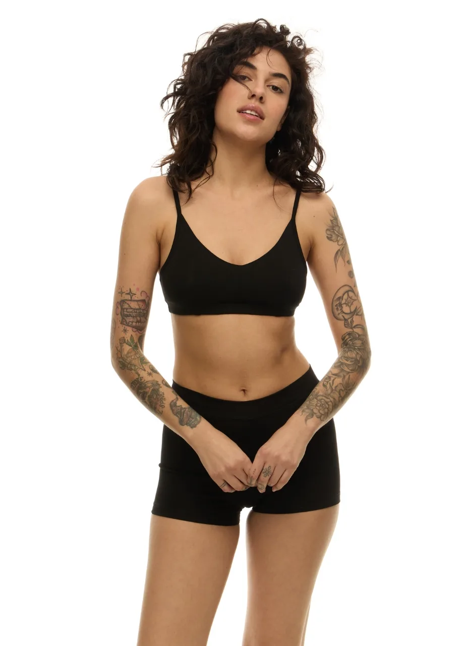 5 › Str/Cd Wear Woman with wavy dark hair wearing a black bralette and matching shorts, tattoos on both arms, posing against a white background.
