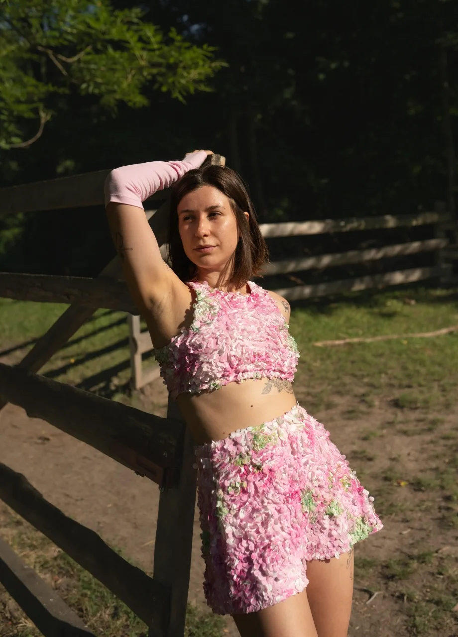 5 › Str/Cd Wear Woman wearing a pink floral two-piece outfit, leaning on a wooden fence outdoors.