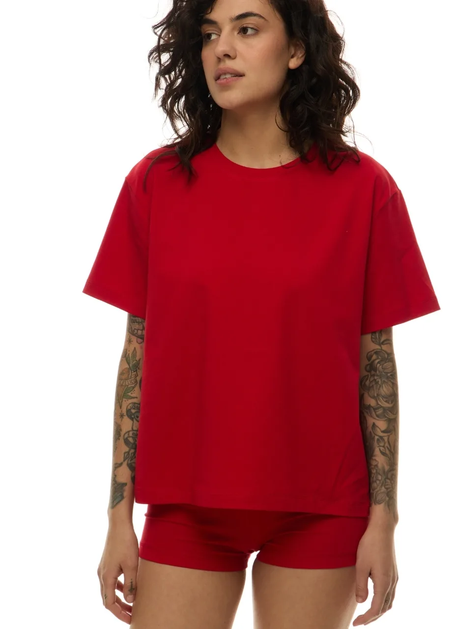 51 › Str/Cd Wear Woman with wavy dark hair wearing a red oversized T-shirt and matching red shorts; tattoos on both forearms visible.
