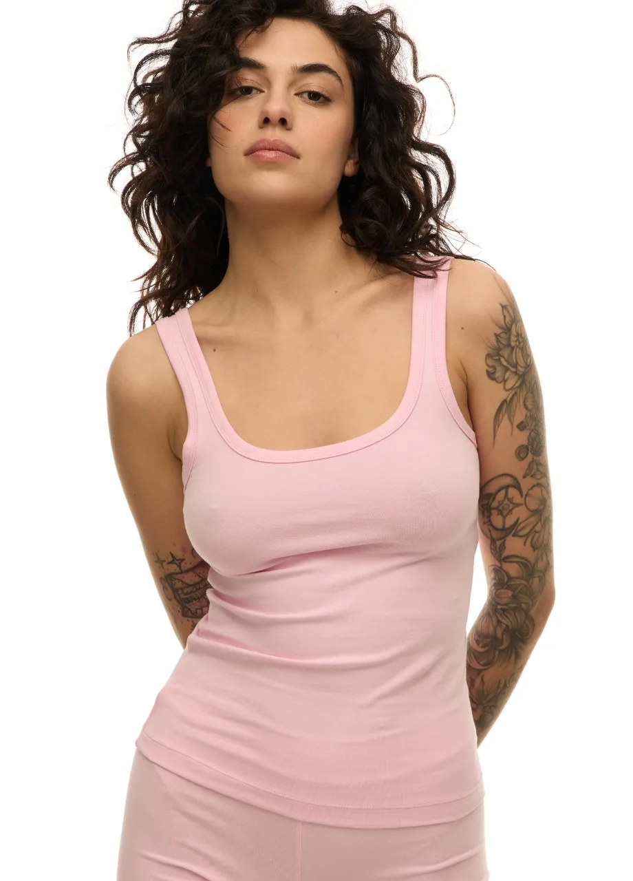 59 › Str/Cd Wear Woman with wavy dark hair wearing a pale pink tank top, standing with arms behind her, tattoos visible on both arms.