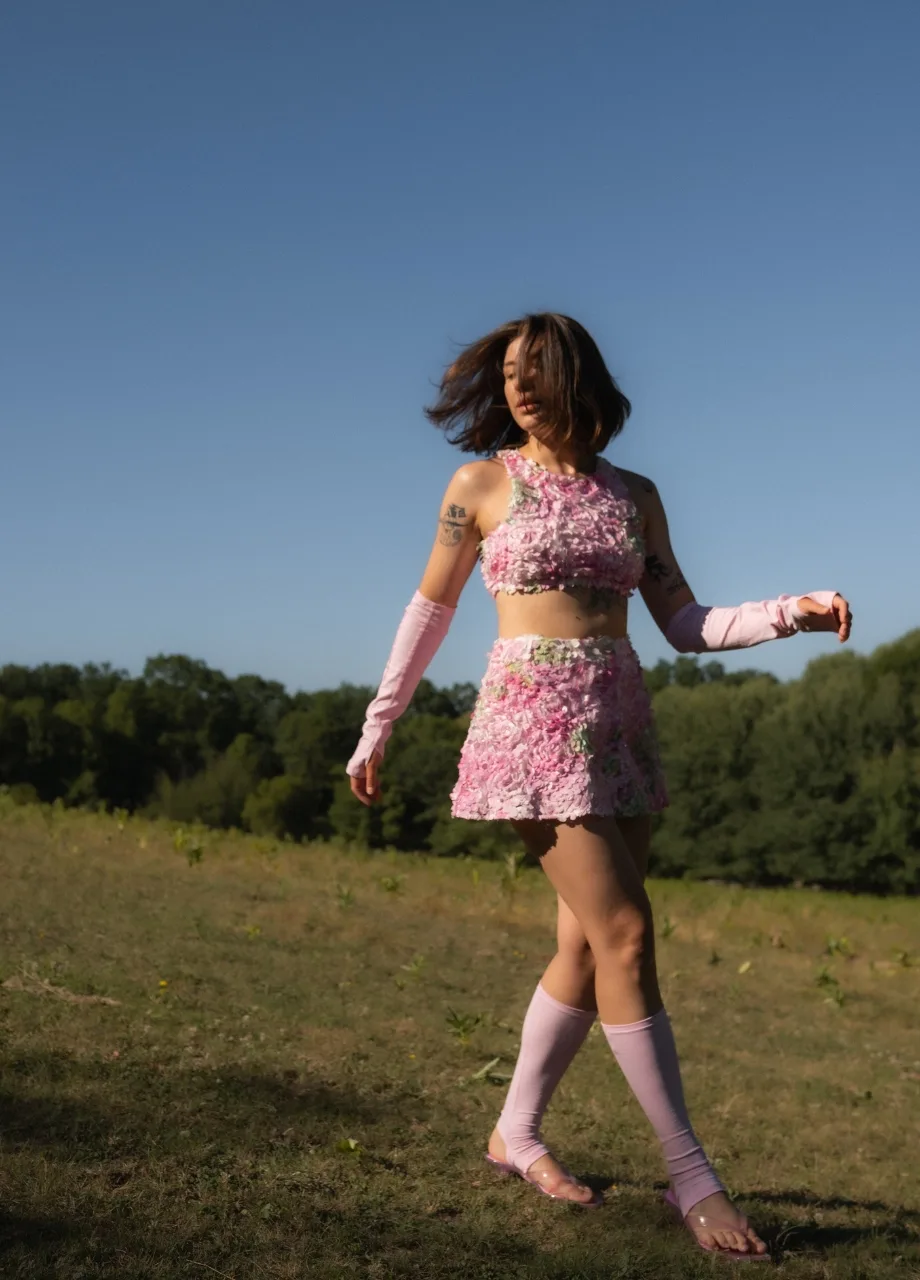 6 › Str/Cd Wear Woman dancing in a pink floral two-piece outfit (crop top and skirt) with matching arm sleeves in a sunlit field, hair blowing in the wind.