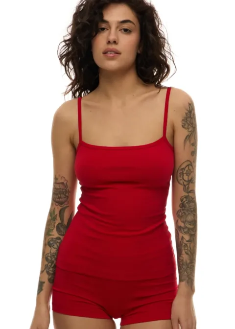 Woman with curly dark hair wearing a red spaghetti-strap tank top and matching red shorts; visible tattoos on both arms against a white background.