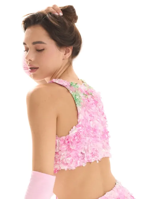 Woman seen from behind wearing a pink, flower-textured crop top with a matching skirt; hair in an updo, head slightly turned downward.