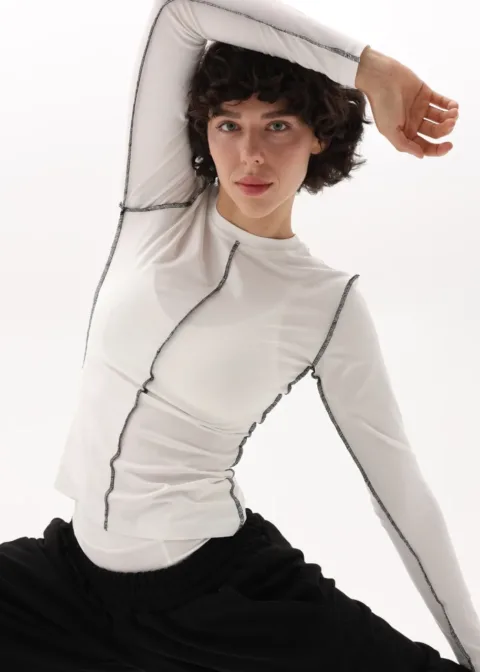 Model wearing a white long-sleeve top with black contrast stitching, posing with arm raised.