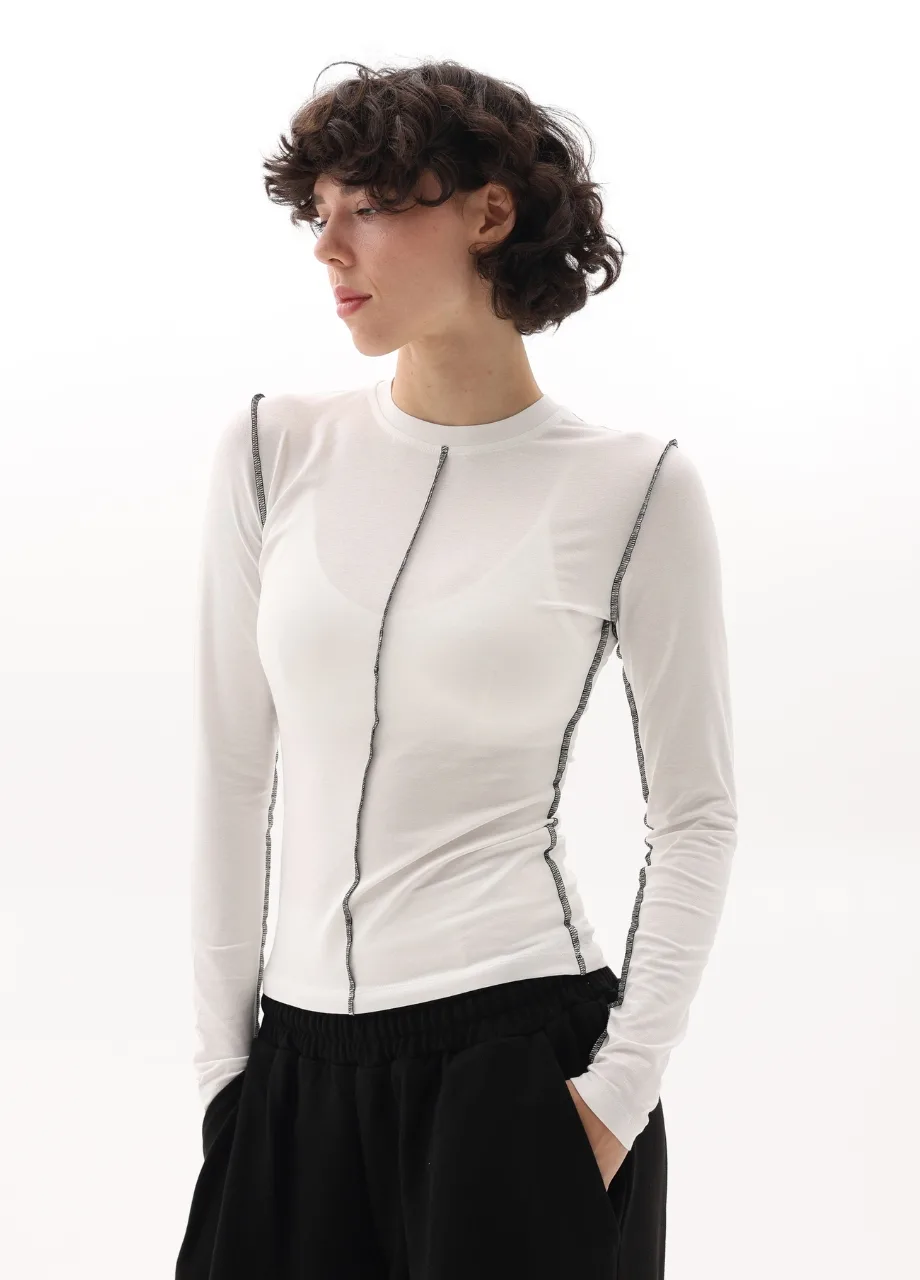 12 › Str/Cd Wear Woman standing in a studio wearing a white long-sleeve top with prominent black stitching seams and black wide-leg pants, hands in pockets, looking to the left