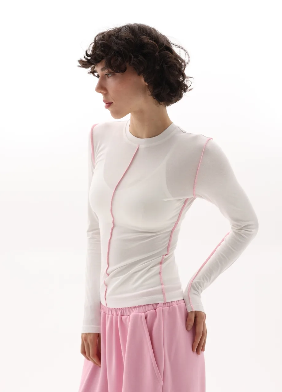 2 › Str/Cd Wear Side-profile of a woman modeling a white long-sleeve top with pink topstitching and a soft pink gathered skirt against a white background.