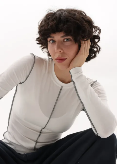 Woman with short curly brown hair wearing a white long-sleeve top with dark stitching, resting her head on her hand in a seated pose against a white background