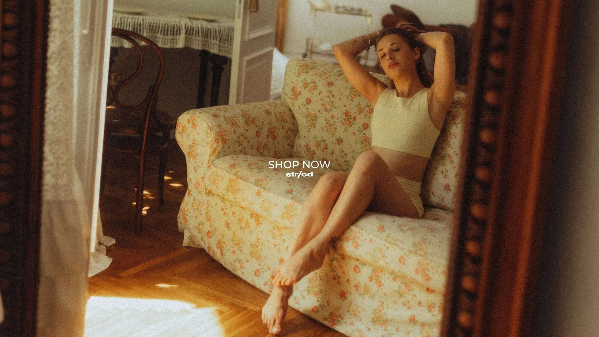 Model lounging on a floral loveseat in a sunlit vintage room, wearing a pale green cropped top and shorts.